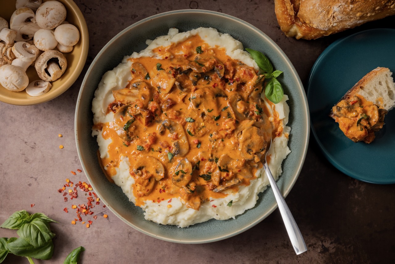 Creamy J-M Farms mushrooms in a tomato-cream sauce tops buttery mashed potatoes, served in a bowl surrounded by the ingredients of the dish.