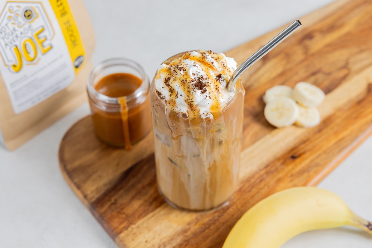 A banana cream cold brew coffee beverage sits on a rustic wood cutting board next to a bag of Not Your Average Joe House Blend coffee beans.