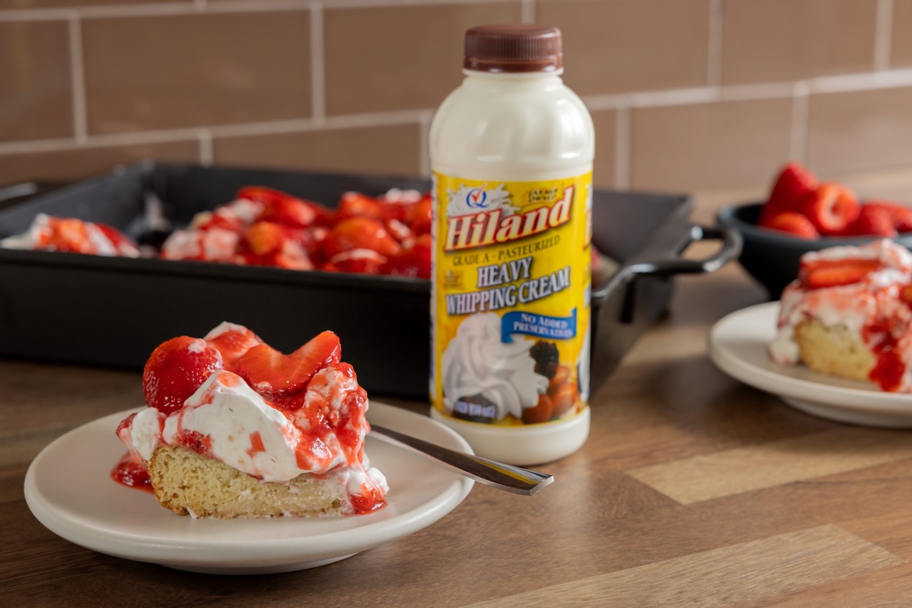 A slice of strawberry shortcake piled high with whipped Hiland Dairy cream sits on a butcher block counter top, with the full strawberry shortcake, large enough to feed a crowd, blurred in the background.