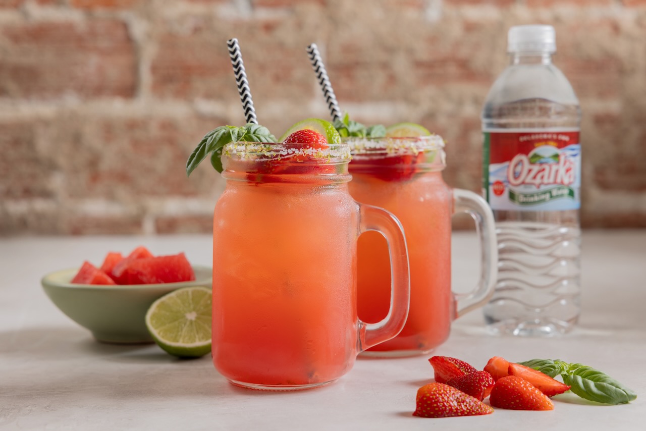 Two mugs of strawberry watermelon agua fresca beverages on a kitchen countertop, with a bottle of Oklahoma's Own Ozarka Drinking Water visible in the background.